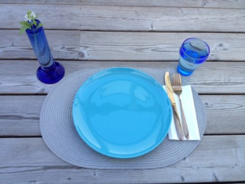 Bildet er under tittelen budsjett mat en person og viser et oppdekt bord for en person med turkis tallerken på sølvgrå bordbrikke med blått glass med vann, serviett og kniv og gaffel samt blomstervase med basilikum blomst i.